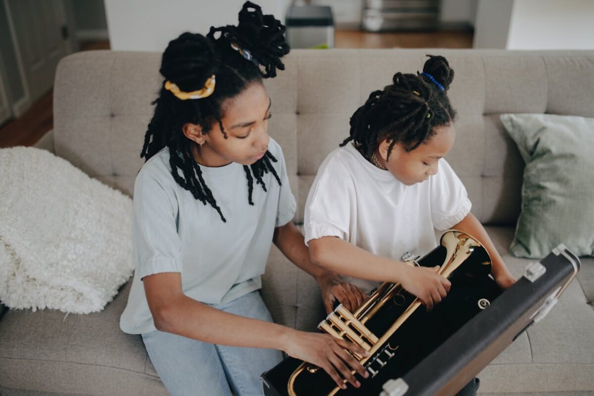 Two young girls with dreadlocks exploring a trumpet together indoors, fostering music interest.